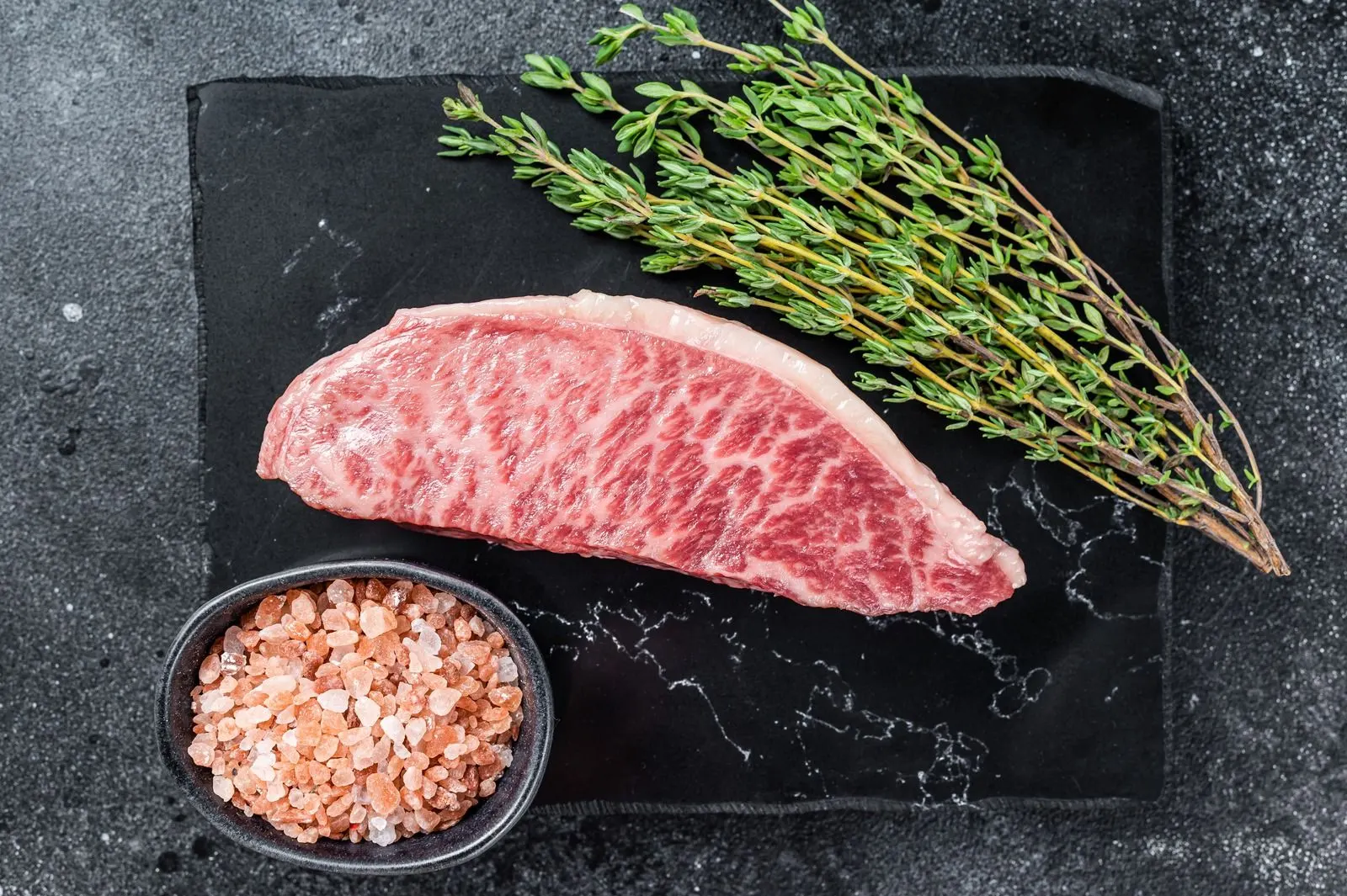 A raw marbled steak on dark slate with thyme and salt